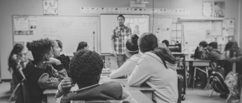 A male teacher standing in front of a class of around 20 teenagers sitting at tables. Photo by Kenny Eliason on Unsplash.