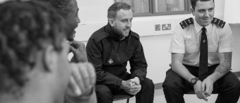 Two male prison officers seated. They are smiling. We can see the backs of one prisoner's head. Another prisoner is shown in profile, laughing.